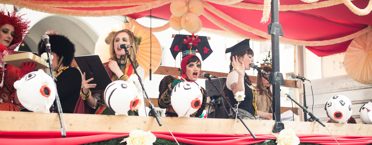 Photo of a float decorated with red fabric, white and green garlands and paper flowers. Several women stand on the float and sing into microphones attached to the float. They are dressed in colourful traditional costumes, wearing headdresses and expressive make-up.