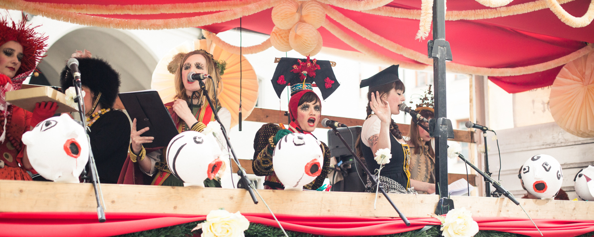 Photo of a float decorated with red fabric, white and green garlands and paper flowers. Several women stand on the float and sing into microphones attached to the float. They are dressed in colourful traditional costumes, wearing headdresses and expressive make-up.
