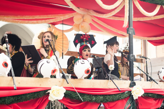 Photo of a float decorated with red fabric, white and green garlands and paper flowers. Several women stand on the float and sing into microphones attached to the float. They are dressed in colourful traditional costumes, wearing headdresses and expressive make-up.