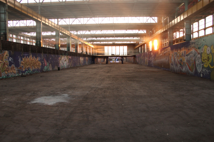 View into the empty industrial hall 24 of the former Freimann railway repair works at sunset. The sun shines through large glass windows, the walls of the hall are painted with graffiti. In the centre of the hall is an installation by artist Emauel Mooner, a colourful neon sign with the words "Today will be a good day".