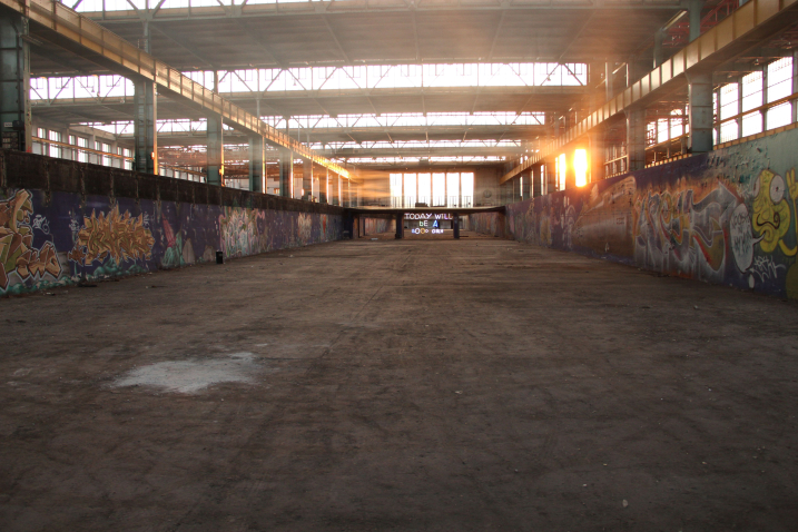 Blick in die leere Industriehalle 24 des ehemaligen Reichsbahn-Ausbesserungswerk Freimann bei Sonnenuntergang. Die Sonne fällt durch großen Glasfenster, die Wände der Halle sind mit Graffiti bemalt. In der Mitte der Halle ist eine Installation des Künstlers Emauel Mooner zu sehen, eine bunte Leuchtschrift mit den Worten "Today will be a good day".