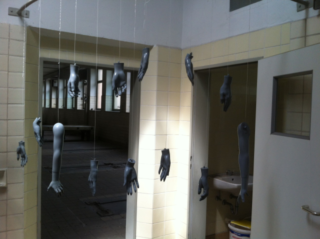 View into a beige-tiled corridor from which two rooms lead off. An open grey door leads into a tiled bathroom with a white washbasin. The second room is a hall with a tiled floor and lots of windows.