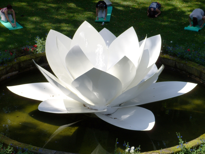 Foto einer Skulptur einer weißen Lotusblüte aus Kunststoff, die mittig in einem von Blumenbeeten umrandeten Wasserbecken platziert ist. Auf der umgebenden Rasenfläche sind einige Menschen auf Yogamatten zu sehen, die eine verbeugende Yogapose vollführen.