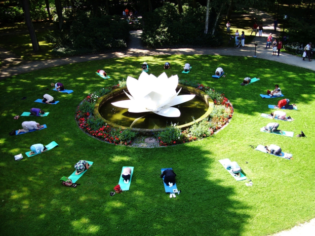 View of the green area of the Marienhof from above. In the centre of the green space is a sculpture of a white plastic lotus flower in a basin of water surrounded by flowerbeds. People on yoga mats are grouped in a circle around the lotus flower on the lawn, performing a bowing yoga pose.