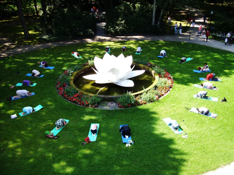 Ansicht der Grünfläche des Marienhofes von oben. In der Mitte der Grünflache zu sehen ist die Skulptur einer weißen Lotusblüte aus Kunststoff in einem von Blumenbeeten umrandeten Wasserbecken. Kreisförmig um die Lotusblüte herum sind Menschen auf Yogamatten auf dem Rasen gruppiert, die eine verbeugende Yogapose vollführen.