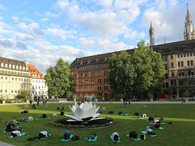 View of the green area of the Marienhof. The back of the New Town Hall can be seen in the background. The sculptur of a white plastic lotus flower is placed in the centre of the green. People on yoga mats are grouped in a circle around the lotus flower on the lawn, performing a bowing yoga pose.