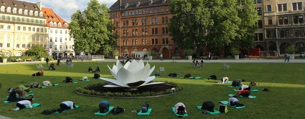 View of the green area of the Marienhof. The back of the New Town Hall can be seen in the background. The sculptur of a white plastic lotus flower is placed in the centre of the green. People on yoga mats are grouped in a circle around the lotus flower on the lawn, performing a bowing yoga pose.