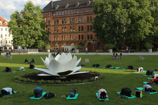 View of the green area of the Marienhof. The back of the New Town Hall can be seen in the background. The sculptur of a white plastic lotus flower is placed in the centre of the green. People on yoga mats are grouped in a circle around the lotus flower on the lawn, performing a bowing yoga pose.