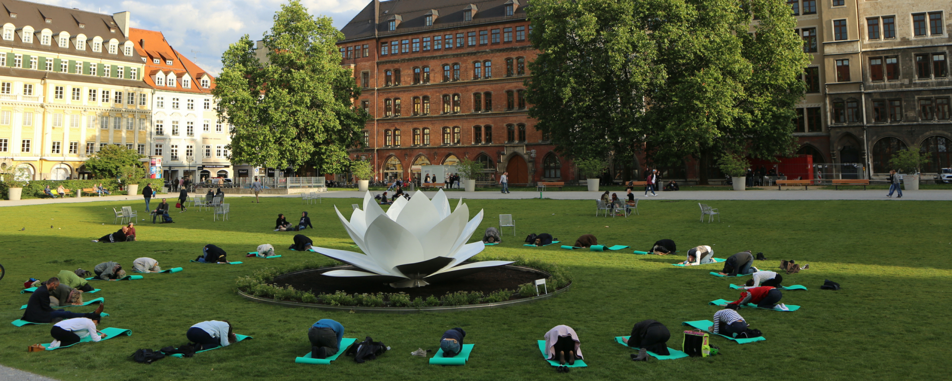 Ansicht der Grünfläche des Marienhofes. Im Hintergrund zu sehen ist die Rückseite des Neuen Rathauses. In der Mitte der Grünflache ist die Skulptur einer weißen Lotusblüte aus Kunststoff platziert. Kreisförmig um die Lotusblüte herum sind Menschen auf Yogamatten auf dem Rasen gruppiert, die eine verbeugende Yogapose vollführen.