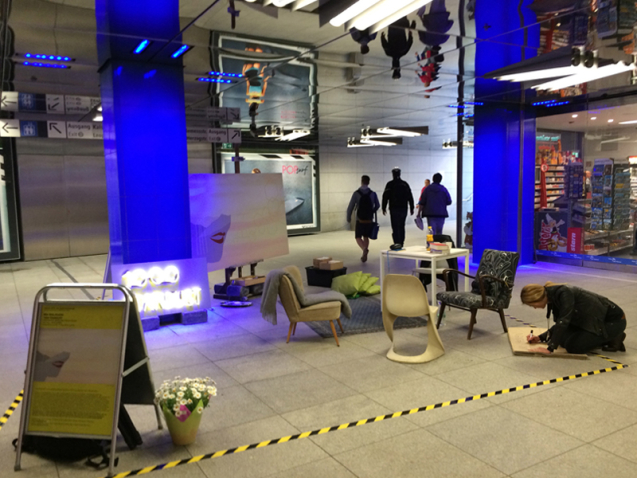 View of the mezzanine level of the Münchner Freiheit underground station. The artist Mia Maljojoki has set up an installation that doubles as a performance space, surrounded by yellow and black tape on the floor. There are four different chairs, a white table, an illuminated billboard with the neon sign "1000 Stardust", a stand with an information text about the project and a flower bed. On a large easel is a rectangular poster. The poster shows a yellow-pink background, reminiscent of a sunrise sky, on which the lower half of a woman's face appears in grey with bright red lips. Golden pentagons, like speech bubbles, protrude from her mouth. A blonde woman sits on the floor, writing with a pen on a piece of cardboard.