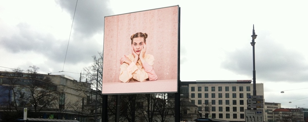 View of the billboard on Lenbachplatz looking towards the city with the motif "Dreiarmige Frau" by Susanne Steinmaßl. The motif shows a blonde woman in a pale pink blouse sitting in front of a pink patterned wallpaper. She has two arms propped up on a pink table in front of her and rests her face in both hands. The woman also has a third arm, which she is resting on the table and clasping her shoulder with her hand.