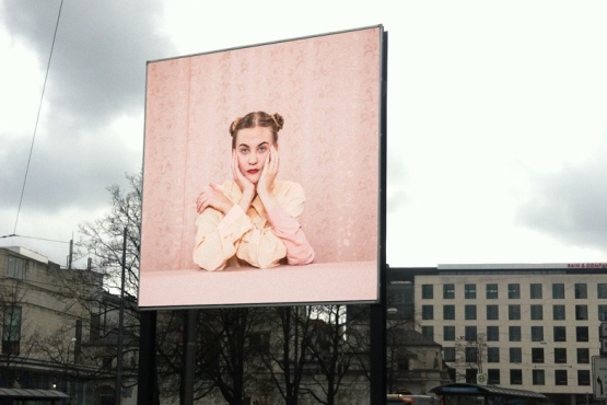 View of the billboard on Lenbachplatz looking towards the city with the motif "Dreiarmige Frau" by Susanne Steinmaßl. The motif shows a blonde woman in a pale pink blouse sitting in front of a pink patterned wallpaper. She has two arms propped up on a pink table in front of her and rests her face in both hands. The woman also has a third arm, which she is resting on the table and clasping her shoulder with her hand.