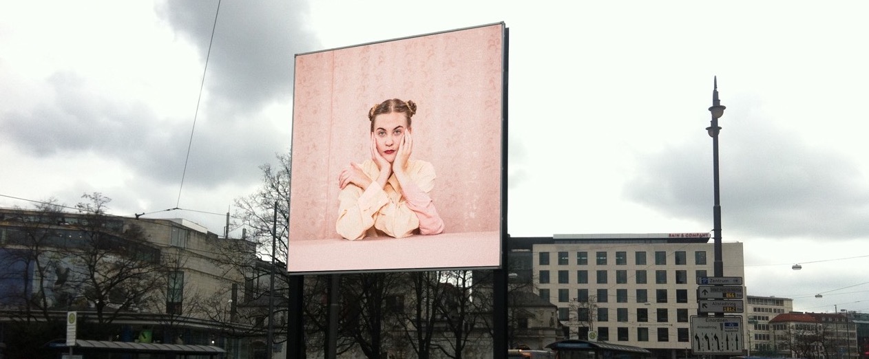 View of the billboard on Lenbachplatz looking towards the city with the motif "Dreiarmige Frau" by Susanne Steinmaßl. The motif shows a blonde woman in a pale pink blouse sitting in front of a pink patterned wallpaper. She has two arms propped up on a pink table in front of her and rests her face in both hands. The woman also has a third arm, which she is resting on the table and clasping her shoulder with her hand.