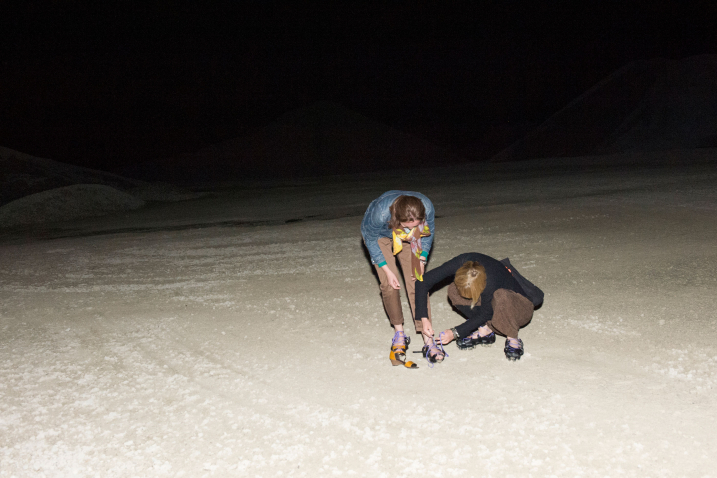 Shot of an illuminated sandy area at night. A woman kneels on the ground and puts a pair of home-made lace-up shoes on another woman. The shoes consist of a black bottom tied together with purple-coloured laces.