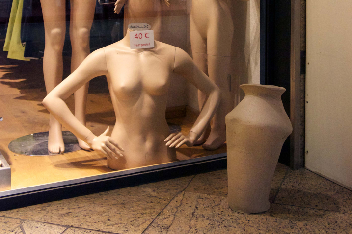 Photograph of a shop window at night. There are four undressed mannequins in the shop window. The front mannequin consists only of a torso without legs and head, on which a price tag with the inscription "40€ fixed price!" is attached. In front of the shop window on the floor is a sculpture that looks like a melted clay amphora.