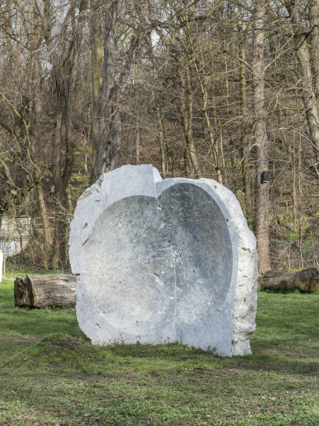 A large grayish stone with a concave hollow stands on a grassy area, with many trees in the background