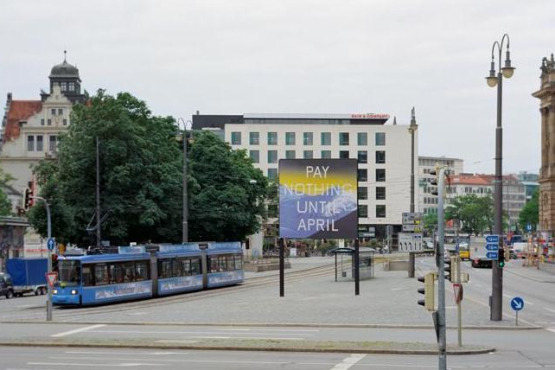 Frontal view of Lenbachplatz looking towards the city center. The billboard is placed in the middle of the square, with a streetcar running alongside. The billboard features a work by the artist Ed Ruscha. The motif shows a snow-covered mountain range in blue with a yellowish horizon above it. The inscription "Pay Nothing Until April" appears on the picture in white capital letters.