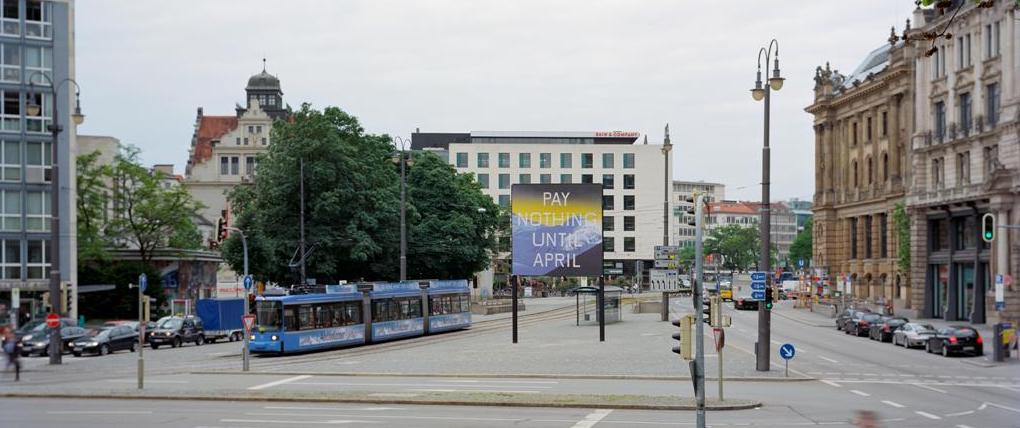 Frontal view of Lenbachplatz looking towards the city center. The billboard is placed in the middle of the square, with a streetcar running alongside. The billboard features a work by the artist Ed Ruscha. The motif shows a snow-covered mountain range in blue with a yellowish horizon above it. The inscription "Pay Nothing Until April" appears on the picture in white capital letters.