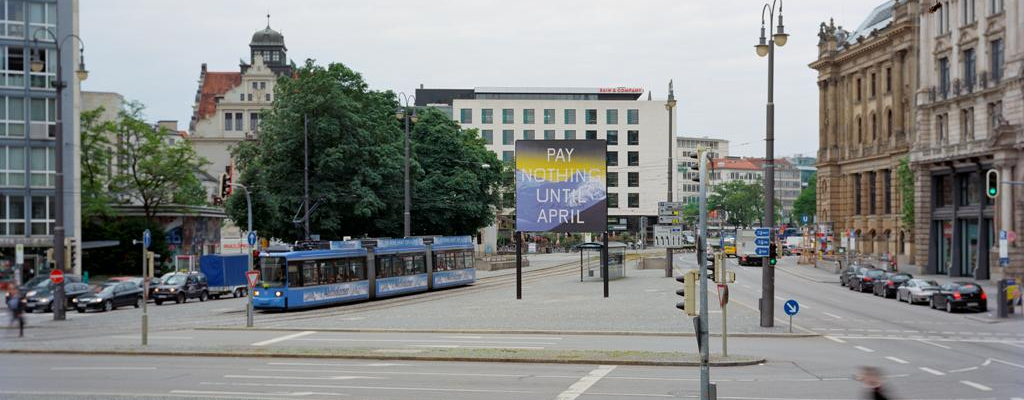 Frontale Ansicht des Lenbachplatzes mit Blick Richtung Innenstadt. In der Mitte der Platzes ist das Billboard platziert, seitlich fährt eine Trambahn dabei. Auf dem Billboard zu sehen ist eine Arbeit des Künstlers Ed Ruscha. Das Motiv zeigt eine in blau gehaltene verschneite Bergkette und darüber ein gelblicher Horizont. Auf dem Bild erscheint die Aufschrift: "Pay Nothing Until April" in weißen Großbuchstaben.