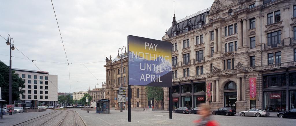 Lateral view of the billboard at Lenbachplatz looking towards the city center. The billboard features a work by the artist Ed Ruscha. The motif shows a snow-covered mountain range in blue with a yellowish horizon above it. The inscription "Pay Nothing Until April" appears on the picture in white capital letters.