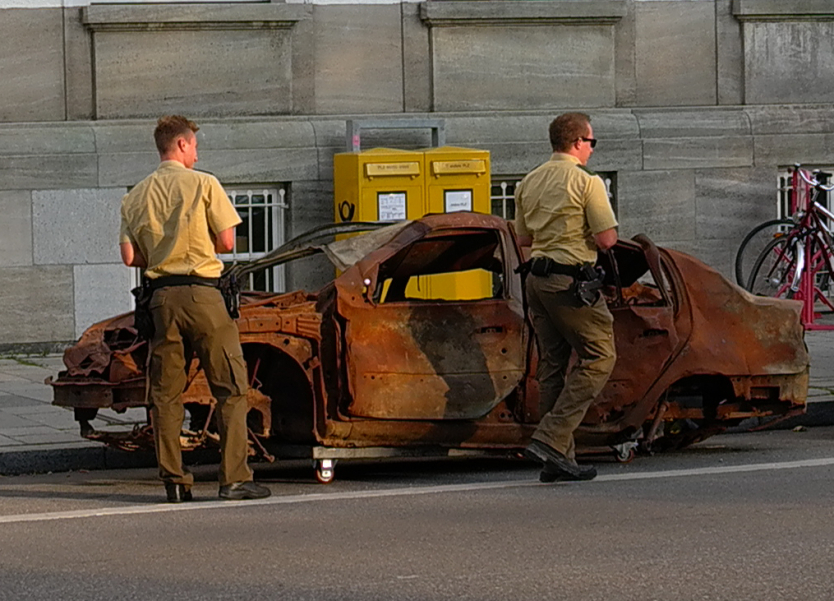 Photograph of a brown, burnt-out car wreck with no tyres, parked on Maximilianstraße in Munich. Two policemen are standing in front of the wreck, walking around the car and inspecting it.
