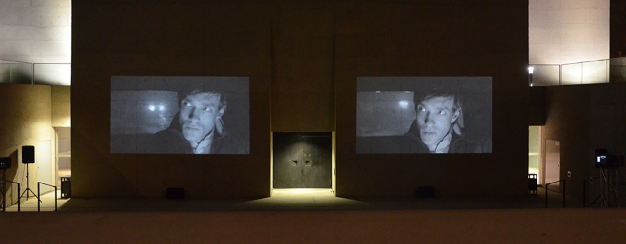 Photograph of a double video projection of the film "Wednesday" by the artist duo M+M on the outer façade of the entrance gate of the State Museum of Egyptian Art. The black and white video stills on display show the same man driving in a car in the dark. His expression is serious and he is looking in the rear-view mirror. The headlights of another car can be seen through the rear window.