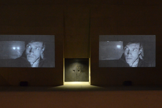 Fotografie einer Doppelvideoprojektion des Films "Mittwoch" des Künstlerduos M+M auf der Außenfassade des Eingangstors des Staatlichen Museums Ägyptischer Kunst. Auf den gezeigten Schwarz-Weiß-Videostills ist der gleiche Mann zu sehen, der in der Dunkelheit in einem Auto fährt. Seine Miene ist ernst und er blickt in den Rückspiegel. Durch die Rückscheibe sind die Scheinwerfer eines weiteren Autos zu sehen.