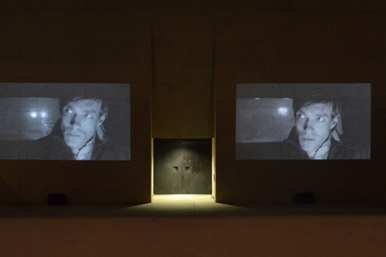 Photograph of a double video projection of the film "Wednesday" by the artist duo M+M on the outer façade of the entrance gate of the State Museum of Egyptian Art. The black and white video stills on display show the same man driving in a car in the dark. His expression is serious and he is looking in the rear-view mirror. The headlights of another car can be seen through the rear window.