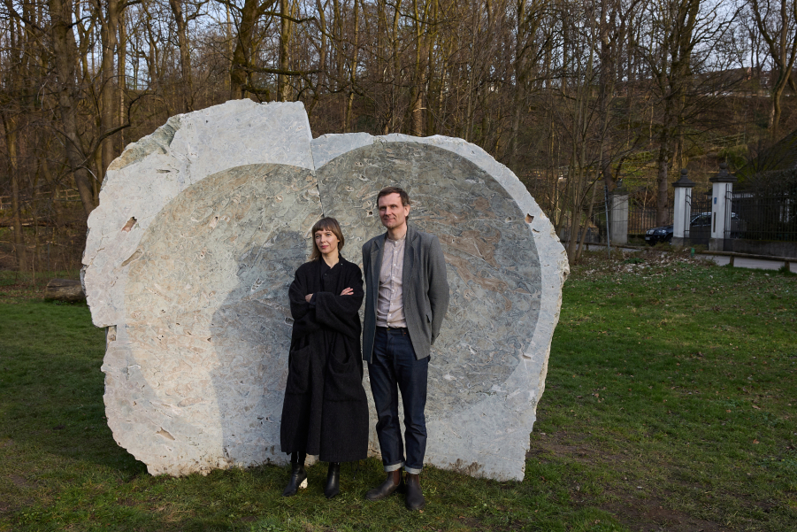 A woman and a man, Lina Lapelytė and Mantas Peteraitis, stand on the grass in front of a large grayish stone with a concave hollow, with many trees in the background