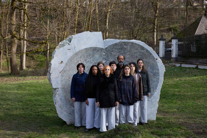 Nine people are standing on a grassy area in front of a large grayish stone with a concave hollow, with many trees in the background