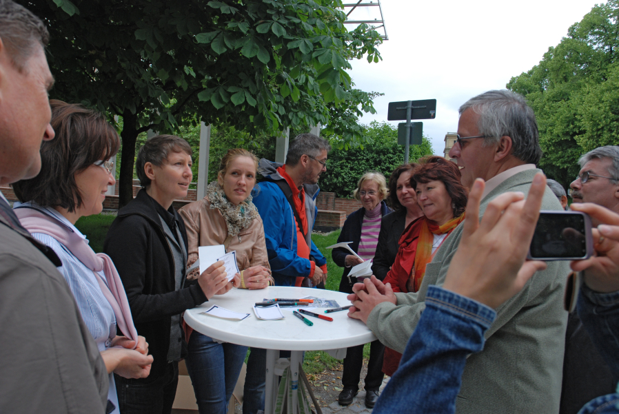 A group of people are standing around a round white bar table and the group is in conversation. There are pens and white and blue pieces of paper on the table. The trees of the courtyard garden can be seen in the background.