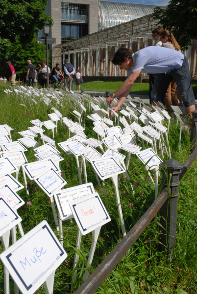 Photograph of a lawn in the Hofgarten with many white signs stuck into it. The signs have words and phrases written on them in different colours. A man can be seen sticking another sign in the grass. Some passers-by on the footpath and the Bavarian State Chancellery can be seen in the background.