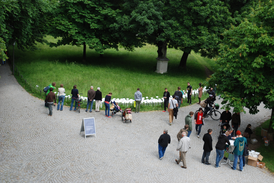 Aerial view of a path in the courtyard garden. You can see a lawn with a large number of white signs stuck in it. A number of passers-by are standing in front of the lawn, reading the signs. There is also a high table on the path with a few people standing around it.