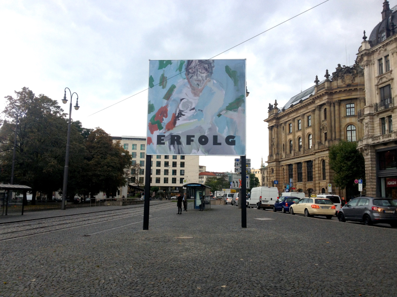 Frontal view of the billboard at Lenbachplatz photographed towards the city centre. The billboard features a work by the artist Stephan Dillemuth. The motif shows an abstract portrait of a naked man against a blue-green background. The word "Erfolg" (success) is written in bold black capital letters in the lower third of the image.