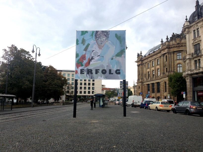 Frontale Ansicht des Billboards am Lenbachplatz in Richtung Innenstadt fotografiert. Auf dem Billboard zu sehen ist eine Arbeit des Künstlers Stephan Dillemuth. Das Motiv zeigt das abstrakt gehaltene Porträt eines nackten Mannes vor einem blau-grünen Hintergrund. Im unteren Bilddrittel steht das Wort "Erfolg" in schwarzen fetten Großbuchstaben.