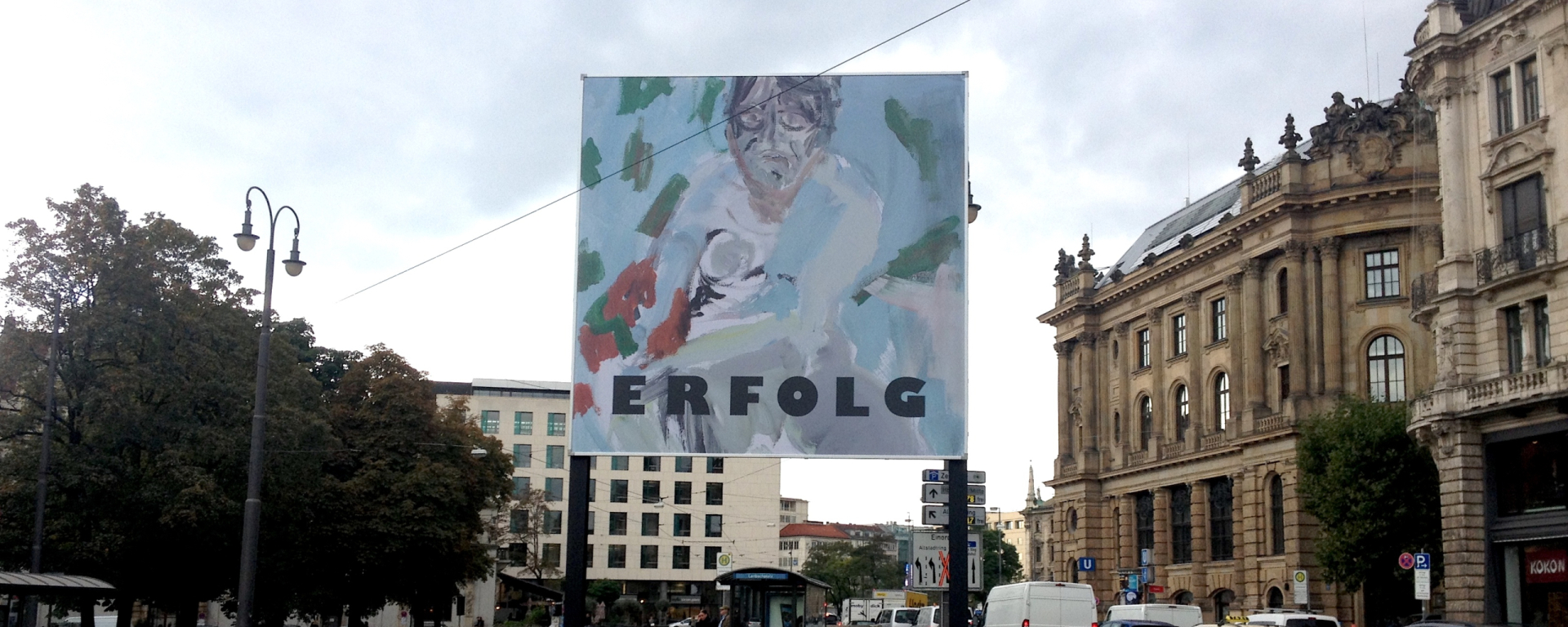 Frontale Ansicht des Billboards am Lenbachplatz in Richtung Innenstadt fotografiert. Auf dem Billboard zu sehen ist eine Arbeit des Künstlers Stephan Dillemuth. Das Motiv zeigt das abstrakt gehaltene Porträt eines nackten Mannes vor einem blau-grünen Hintergrund. Im unteren Bilddrittel steht das Wort "Erfolg" in schwarzen fetten Großbuchstaben.