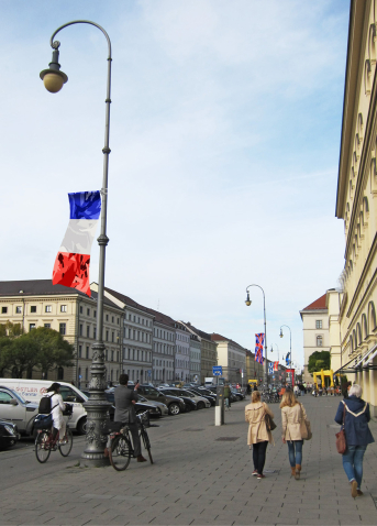 Ansicht der Ludwigstraße mit Passant*innen und Radfahrer*innen vom Odeonsplatz aus gesehen. An den Straßenlaternen sind im Zuge einer Kunstinstallation der Künstlerin Silke Witzsch auf halber Höhe die Nationalflaggen verschiedener Länder angebracht. Die Flaggen sind künstlerisch verfremdet, indem ein Tarnmuster über die Nationalfahnen gelegt wird.