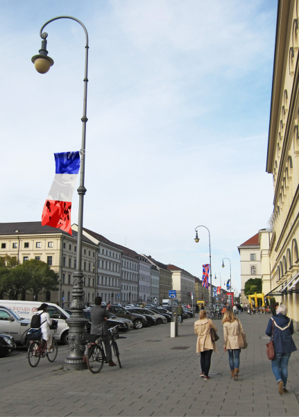 View of Ludwigstraße with pedestrians and cyclists seen from Odeonsplatz. As part of an art installation by artist Silke Witzsch, the national flags of various countries are attached to the street lamps at half height. The flags are artistically distorted by placing a camouflage pattern over the national flags.