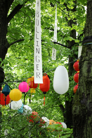 Fotografie der Bäume am Platz der Freiheit in München-Neuhausen. In den Bäumen hängen bunte leuchtende Lampions sowie weiße lange Banner mit den Namen Münchner Widerstandskämpfer*innen in schwarzen Großbuchstaben.