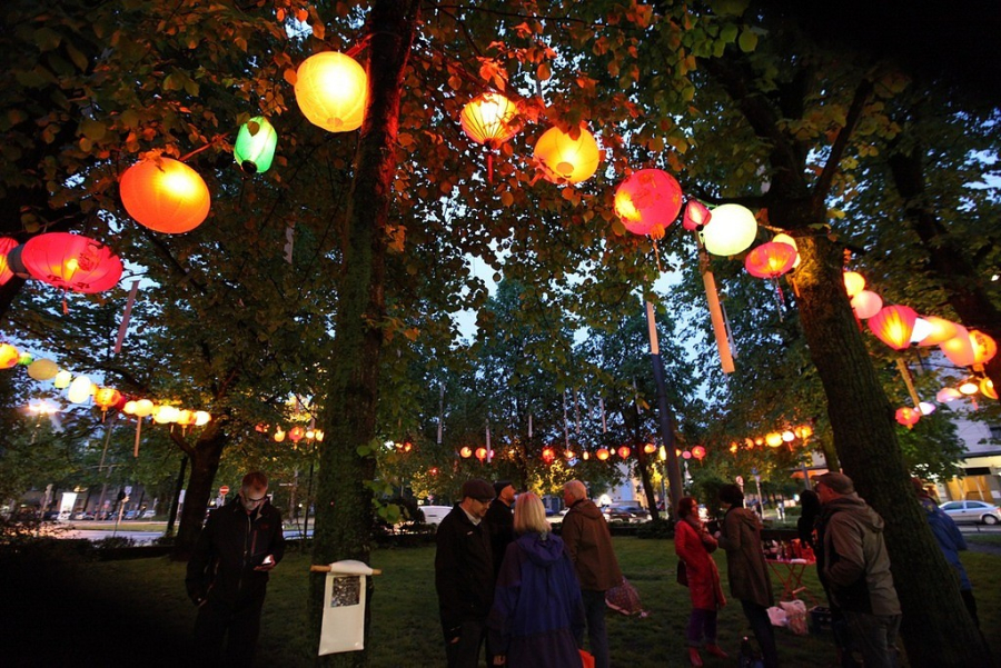 Photograph of the Platz der Freiheit in Munich-Neuhausen in the evening. Brightly coloured lanterns and long white banners hang in the trees on the square. Groups of people are standing on the lawn of the square, chatting.
