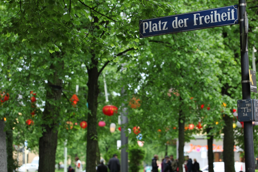 Fotografie der Bäume am Platz der Freiheit in München-Neuhausen. Im Bildvordergrund zu sehen das blaue Straßenschild mit "Platz der Freiheit". Dahinter die Bäume, in denen bunte leuchtende Lampions hängen sowie eine Gruppe von Menschen, die unscharf im Hintergrund zu sehen sind.