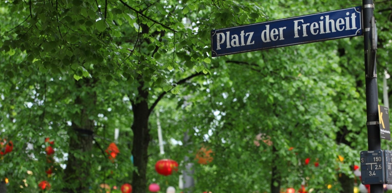 Fotografie der Bäume am Platz der Freiheit in München-Neuhausen. Im Bildvordergrund zu sehen das blaue Straßenschild mit "Platz der Freiheit". Dahinter die Bäume, in denen bunte leuchtende Lampions hängen.