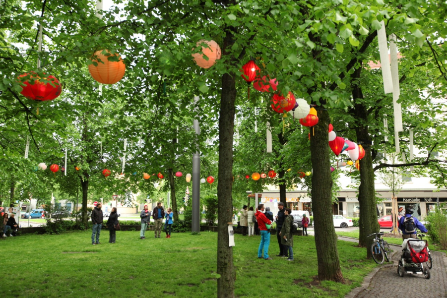 Fotografie des Platzes der Freiheit in München-Neuhausen. In den Bäumen am Platz hängen bunte leuchtende Lampions sowie weiße lange Banner. Auf der Wiese des Platzes stehen Gruppen von Personen, die sich unterhalten.