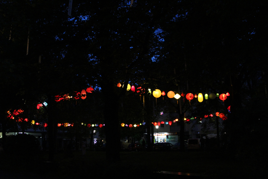 Fotografie des Platzes der Freiheit in München-Neuhausen am Abend. In den Bäumen am Platz hängen bunte leuchtende Lampions sowie weiße lange Banner.