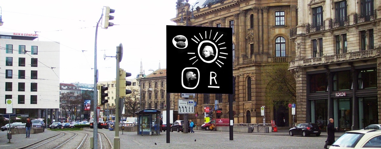 Seitliche Ansicht des Billboards am Lenbachplatz mit Blick Richtung Innenstadt. Auf dem Billboard zu sehen ist eine Arbeit des Künstlers Martin Fengel. Das Motiv zeigt auf schwarzem Hintergrund vier Objekte. Ein weißes unterstrichenes "R" in Großbuchstaben. Ein weißer Kreis mit einem grauen Bild von zwei Kartoffeln. Ein weißer Kreis mit seitlich ausgehenden Strahlen, in dessen Mitte ein Schwarz-Weiß-Porträt des Grafen Rumfords sitzt. Ein zweiter Kringel mit einem Schwarz-Weiß-Porträt eines Mannes im Profil, eventuell ebenfalls Graf Rumford.