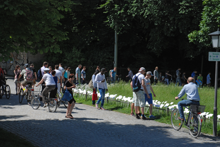 Photograph of a path in the courtyard garden. It shows a lawn with a large number of white signs stuck in it. A number of passers-by are standing in front of the lawn, reading the signs.