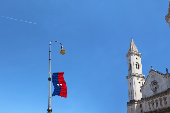 Ansicht einer Straßenlaterne auf der Ludwigstraße von unten fotografiert. An der Straßenlaterne ist im Zuge einer Kunstinstallation der Künstlerin Silke Witzsch auf halber Höhe die Nationalflagge Haitis angebracht. Die Flagge ist künstlerisch verfremdet, indem ein Tarnmuster über die Nationalfahne gelegt wird.