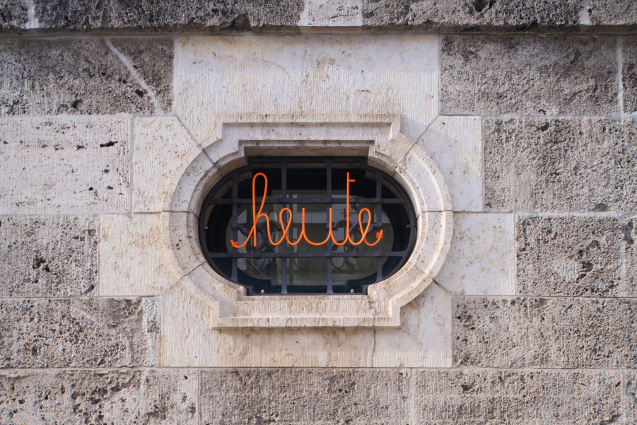 The image shows a house wall with a window grille. The grille features a swastika pattern, and in front of it is the curved orange lettering "today."
