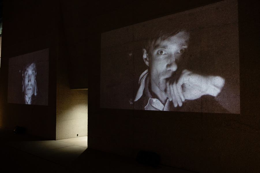 Photograph of a double video projection of the film "Wednesday" by the artist duo M+M on the outer façade of the entrance gate of the State Museum of Egyptian Art. The black and white video stills on display show the same man in frontal view, driving in a car in the dark. His expression is serious and he is leaning his face on his hand, which is resting on the armrest.