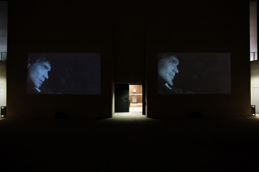 Fotografie einer Doppelvideoprojektion des Films "Mittwoch" des Künstlerduos M+M auf der Außenfassade des Eingangstors des Staatlichen Museums Ägyptischer Kunst. Auf den gezeigten Schwarz-Weiß-Videostills ist der gleiche Mann in Profilansicht zu sehen, der in der Dunkelheit in einem Auto fährt. Seine Miene ist ernst.
